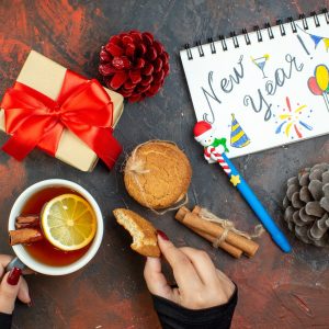 top view cup of tea and cookie in female hand xmas gift pinecones cinnamon sticks cookies new year written on notepad on dark red table