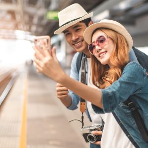 Happy Asian couple traveler holding a mobile phone in station and waiting for train in vacation time.Two Asian Tourists With Backpacks Train travel in Sightseeing City Thailand.