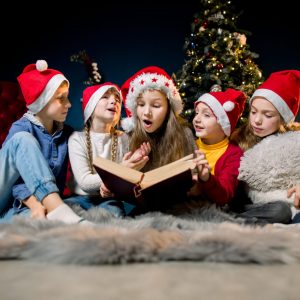 Charming and beautiful little children in Santa's hats are reading Christmas stories and laughing, on the background of a Christmas tree