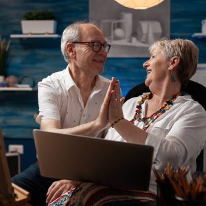 Elderly artist couple holding laptop touching hands showing affection celebrating online success sitting in home studio. Senior art creators enjoying positive feedback on social media.
