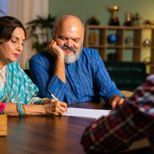 Asian Indian elderly couple carefully signing an important document at home or in a cozy office, sitting across an official at a table, focusing on paperwork and decisions