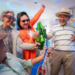 Group of Asian senior friend having evening party in living room at home. Mature elderly man and woman having fun, dancing with cool music while celebrate weekend reunion gathered together in house.