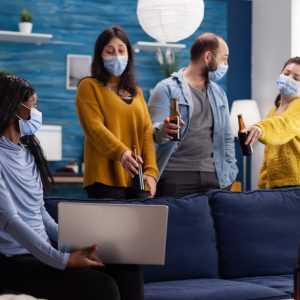 Group of cheerful happy multi ethnic friends looking at photo on laptop while having fun together, chatting keeping social distancing wearing face mask, drinking beer in living room. Conceptual image.
