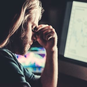Handsome young businessman with shoulder-length blond hair is massaging nose bridge while working with a computer at night