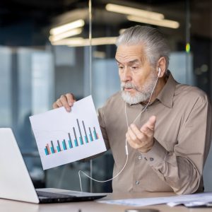 Senior businessman holding a chart during an online video conference in a modern office setting, discussing business data and analytics.