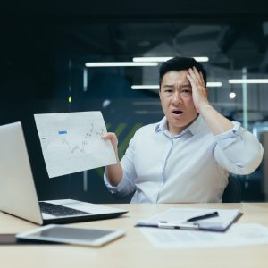 Shocked asian financier showing document with graph with negative figures to camera, man working in office, businessman disappointed with result
