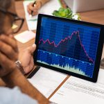 Closeup of a stock market broker working with graphs on digital tablet at office. Rear view of stock agent reading bad report and graph. Back view of multiethnic businessman analyzing fall sales.
