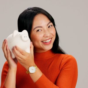 Woman, happy and listen to piggy bank in studio with laughing, saving and coins by background. Japa.